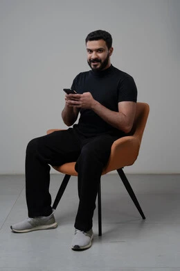 استخدام التقنية الحديثه المتطورة في الحياة العملية، العمل عن بعد عبر الجوال، ايماءات الاندماج والتركيز، بورتريه لشاب عربي خليجي سعودي يرتدي الزي الكاجوال يجلس على الكرسي يقوم بالعمل على الهاتف المحمول، خلفية رمادية