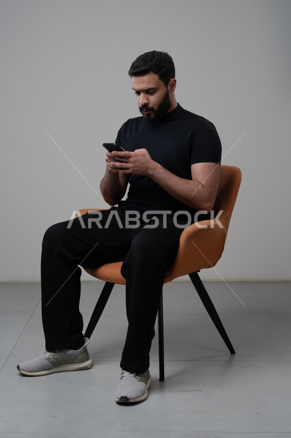 Using modern advanced technology in practical life, working remotely via mobile, gestures of integration and concentration, portrait of a young Saudi Arabian Gulf man wearing casual clothes sitting on a chair working on a mobile phone, gray background