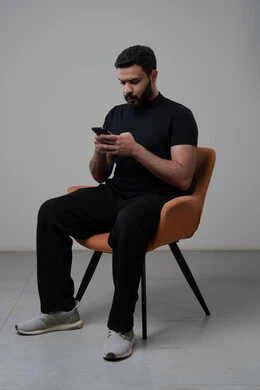 Using modern advanced technology in practical life, working remotely via mobile, gestures of integration and concentration, portrait of a young Saudi Arabian Gulf man wearing casual clothes sitting on a chair working on a mobile phone, gray background