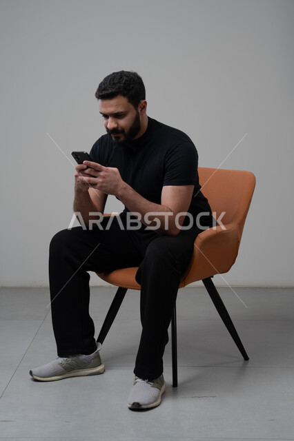 Remote work via mobile, gestures of integration and focus, use of modern advanced technology in practical life, portrait of a young Saudi Arabian Gulf man wearing casual clothes sitting on a chair working on a mobile phone, gray background