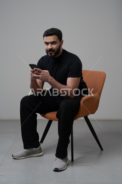 العمل عن بعد عبر الجوال، ايماءات الاندماج والتركيز، استخدام التقنية الحديثه المتطورة في الحياة العملية، بورتريه لشاب عربي خليجي سعودي يرتدي الزي الكاجوال يجلس على الكرسي يقوم بالعمل على الهاتف المحمول، خلفية رمادية