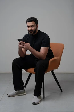 العمل عن بعد عبر الجوال، ايماءات الاندماج والتركيز، استخدام التقنية الحديثه المتطورة في الحياة العملية، بورتريه لشاب عربي خليجي سعودي يرتدي الزي الكاجوال يجلس على الكرسي يقوم بالعمل على الهاتف المحمول، خلفية رمادية