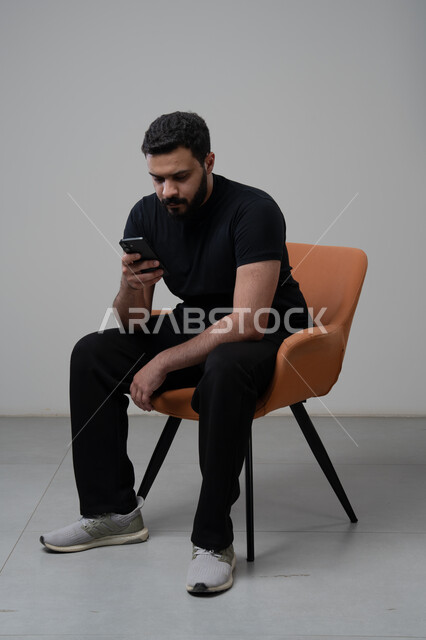العمل عن بعد عبر الجوال، ايماءات الاندماج والتركيز، استخدام التقنية الحديثه المتطورة في الحياة العملية، بورتريه لشاب عربي خليجي سعودي يرتدي الزي الكاجوال يجلس على الكرسي يقوم بالعمل على الهاتف المحمول، خلفية رمادية