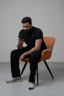 Remote work via mobile, gestures of integration and focus, use of modern advanced technology in practical life, portrait of a young Saudi Arabian Gulf man wearing casual clothes sitting on a chair working on a mobile phone, gray background