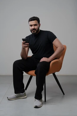 العمل عن بعد عبر الجوال، ايماءات الاندماج والتركيز، استخدام التقنية الحديثه المتطورة في الحياة العملية، بورتريه لشاب عربي خليجي سعودي يرتدي الزي الكاجوال يجلس على الكرسي يقوم بالعمل على الهاتف المحمول، خلفية رمادية