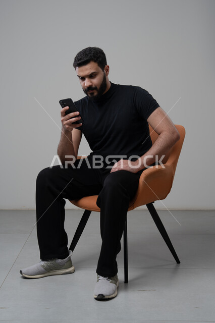 العمل عن بعد عبر الجوال، ايماءات الاندماج والتركيز، استخدام التقنية الحديثه المتطورة في الحياة العملية، بورتريه لشاب عربي خليجي سعودي يرتدي الزي الكاجوال يجلس على الكرسي يقوم بالعمل على الهاتف المحمول، خلفية رمادية
