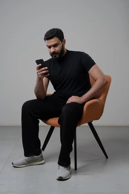 العمل عن بعد عبر الجوال، ايماءات الاندماج والتركيز، استخدام التقنية الحديثه المتطورة في الحياة العملية، بورتريه لشاب عربي خليجي سعودي يرتدي الزي الكاجوال يجلس على الكرسي يقوم بالعمل على الهاتف المحمول، خلفية رمادية