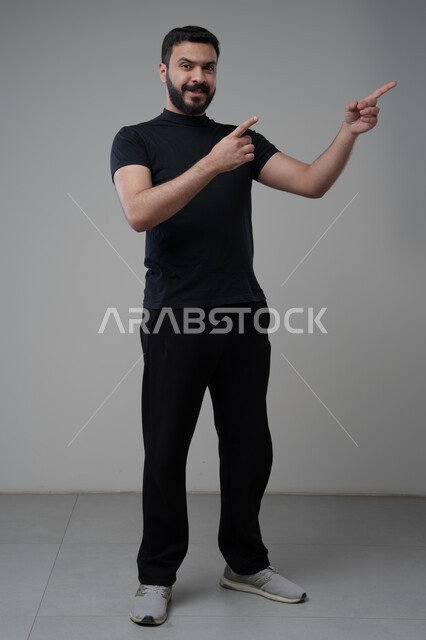 الإشارة إلى شيء ما, الوقوف باستقامة واعتدال, بورتريه لرجل عربي خليجي سعودي يرتدي الزي الكاجوال يشير باصبعي السبابة لمكان معين, صورة للجسم بالطول الكامل, خلفية رمادية