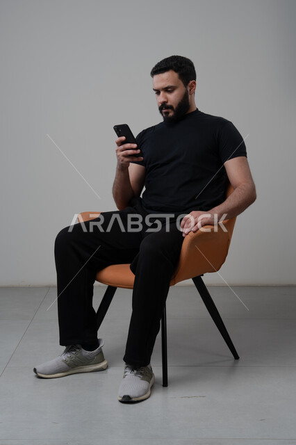 العمل عن بعد عبر الجوال، ايماءات الاندماج والتركيز، استخدام التقنية الحديثه المتطورة في الحياة العملية، بورتريه لشاب عربي خليجي سعودي يرتدي الزي الكاجوال يجلس على الكرسي يقوم بالعمل على الهاتف المحمول، خلفية رمادية