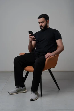 العمل عن بعد عبر الجوال، ايماءات الاندماج والتركيز، استخدام التقنية الحديثه المتطورة في الحياة العملية، بورتريه لشاب عربي خليجي سعودي يرتدي الزي الكاجوال يجلس على الكرسي يقوم بالعمل على الهاتف المحمول، خلفية رمادية