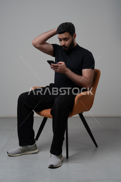 ايماءات الاندماج والتركيز، استخدام التقنية الحديثه المتطورة في الحياة العملية، العمل عن بعد عبر الجوال، بورتريه لشاب عربي خليجي سعودي يرتدي الزي الكاجوال يضع يده على رأسه بايماءات الذهول ويعمل على الهاتف المحمول، خلفية رمادية