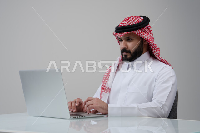 Office profession and job, remote work, use of modern advanced technology in practical life, gestures of integration and focus, portrait of a Saudi Arabian Gulf man wearing traditional thobe and shemagh working on a laptop at company headquarters, white background