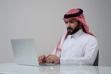 مهنة ووظيفة مكتبية، العمل عن بعد، استخدام التقنية الحديثه المتطورة في الحياة العملية، ايماءات الاندماج والتركيز، بورتريه لرجل عربي خليجي سعودي يرتدي الثوب التقليدي والشماغ يقوم بالعمل على الحاسب المحمول في مقر الشركة، خلفية بيضاء