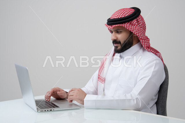مهنة ووظيفة مكتبية، العمل عن بعد، استخدام التقنية الحديثه المتطورة في الحياة العملية، ايماءات الاندماج والتركيز، بورتريه لرجل عربي خليجي سعودي يرتدي الثوب التقليدي والشماغ يقوم بالعمل على الحاسب المحمول في مقر الشركة، خلفية بيضاء