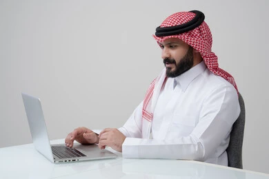 مهنة ووظيفة مكتبية، العمل عن بعد، استخدام التقنية الحديثه المتطورة في الحياة العملية، ايماءات الاندماج والتركيز، بورتريه لرجل عربي خليجي سعودي يرتدي الثوب التقليدي والشماغ يقوم بالعمل على الحاسب المحمول في مقر الشركة، خلفية بيضاء