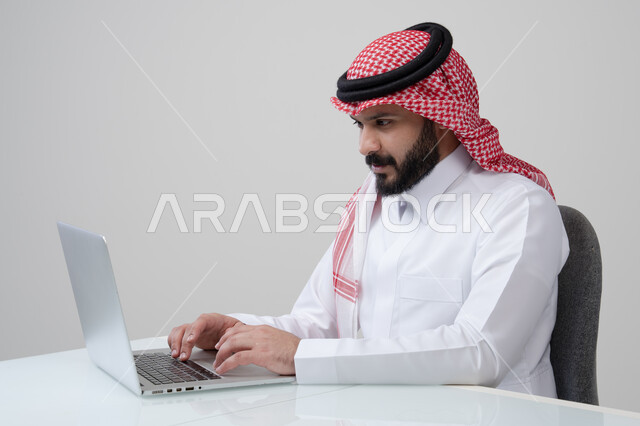مهنة ووظيفة مكتبية، العمل عن بعد، استخدام التقنية الحديثه المتطورة في الحياة العملية، ايماءات الاندماج والتركيز، بورتريه لرجل عربي خليجي سعودي يرتدي الثوب التقليدي والشماغ يقوم بالعمل على الحاسب المحمول في مقر الشركة، خلفية بيضاء