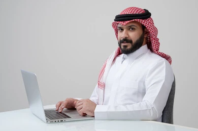 مهنة ووظيفة مكتبية، العمل عن بعد، استخدام التقنية الحديثه المتطورة في الحياة العملية، ايماءات الاندماج والتركيز، بورتريه لرجل عربي خليجي سعودي يرتدي الثوب التقليدي والشماغ يقوم بالعمل على الحاسب المحمول في مقر الشركة، خلفية بيضاء