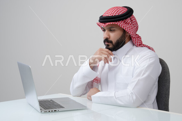مهنة ووظيفة مكتبية، العمل عن بعد، استخدام التقنية الحديثه المتطورة في الحياة العملية، ايماءات الاندماج والتركيز، بورتريه لرجل عربي خليجي سعودي يرتدي الثوب التقليدي والشماغ يقوم بالعمل على الحاسب المحمول في مقر الشركة، خلفية بيضاء