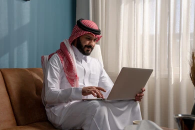 Remote work, office career and job, use of modern advanced technology in practical life, gestures of integration and focus, a Saudi Arabian Gulf man wearing traditional thobe and shemagh sitting on the sofa and working on the laptop in the living room