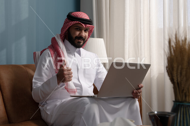 العمل عن بعد، مهنة ووظيفة مكتبية، استخدام التقنية الحديثه المتطورة في الحياة العملية، ايماءات الاندماج والتركيز، رجل عربي خليجي سعودي يرتدي الثوب التقليدي والشماغ يجلس على الكنبة ويعمل على الحاسب المحمول في صالة المنزل