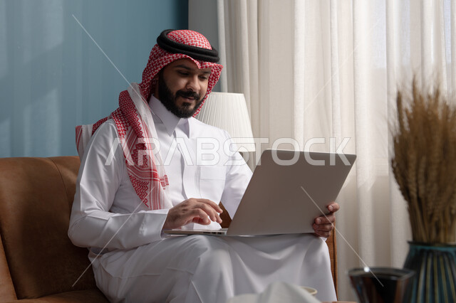العمل عن بعد، مهنة ووظيفة مكتبية، استخدام التقنية الحديثه المتطورة في الحياة العملية، ايماءات الاندماج والتركيز، رجل عربي خليجي سعودي يرتدي الثوب التقليدي والشماغ يجلس على الكنبة ويعمل على الحاسب المحمول في صالة المنزل