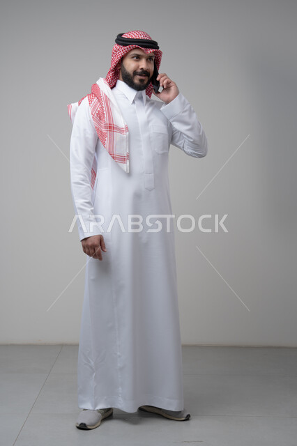 Chatting and conversation using a modern technical device, answering a phone call, portrait of a Saudi Arabian Gulf man wearing traditional thobe making a phone call through a mobile phone, full body image, white background