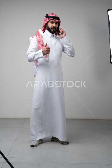 Gestures of approval and satisfaction, raising the thumbs up as an expression of approval, chatting and conversing using a modern technical device, answering a phone call, portrait of a Saudi Arabian Gulf man wearing traditional thobe making a phone call using a mobile phone, full body, white background