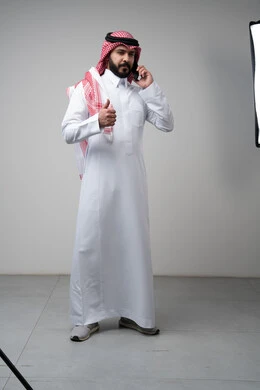 Gestures of approval and satisfaction, raising the thumbs up as an expression of approval, chatting and conversing using a modern technical device, answering a phone call, portrait of a Saudi Arabian Gulf man wearing traditional thobe making a phone call using a mobile phone, full body, white background