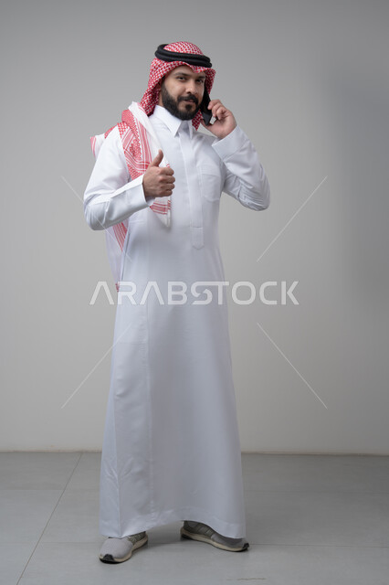 Gestures of approval and satisfaction, raising the thumbs up as an expression of approval, chatting and conversing using a modern technical device, answering a phone call, portrait of a Saudi Arabian Gulf man wearing traditional thobe making a phone call using a mobile phone, full body, white background