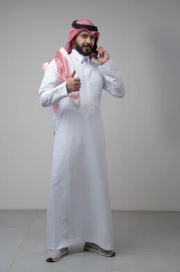 Gestures of approval and satisfaction, raising the thumbs up as an expression of approval, chatting and conversing using a modern technical device, answering a phone call, portrait of a Saudi Arabian Gulf man wearing traditional thobe making a phone call using a mobile phone, full body, white background