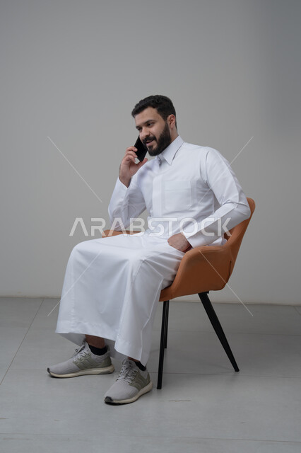 الرد على مكالمة هاتفية عبر الجوال، الدردشة والمحادثة باستخدام جهاز تقني حديث, بورتريه لرجل عربي خليجي سعودي يرتدي الثوب التقليدي يجلس على الكرسي يقوم باجراء اتصال هاتفي من خلال الهاتف المحمول، خلفية بيضاء