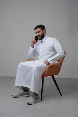 Answering a phone call via mobile phone, chatting and talking using a modern technical device, portrait of a Saudi Arabian Gulf man wearing traditional thobe sitting on a chair making a phone call via mobile phone, white background
