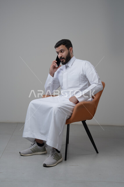 الرد على مكالمة هاتفية عبر الجوال، الدردشة والمحادثة باستخدام جهاز تقني حديث, بورتريه لرجل عربي خليجي سعودي يرتدي الثوب التقليدي يجلس على الكرسي يقوم باجراء اتصال هاتفي من خلال الهاتف المحمول، خلفية بيضاء