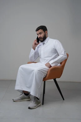 الرد على مكالمة هاتفية عبر الجوال، الدردشة والمحادثة باستخدام جهاز تقني حديث, بورتريه لرجل عربي خليجي سعودي يرتدي الثوب التقليدي يجلس على الكرسي يقوم باجراء اتصال هاتفي من خلال الهاتف المحمول، خلفية بيضاء