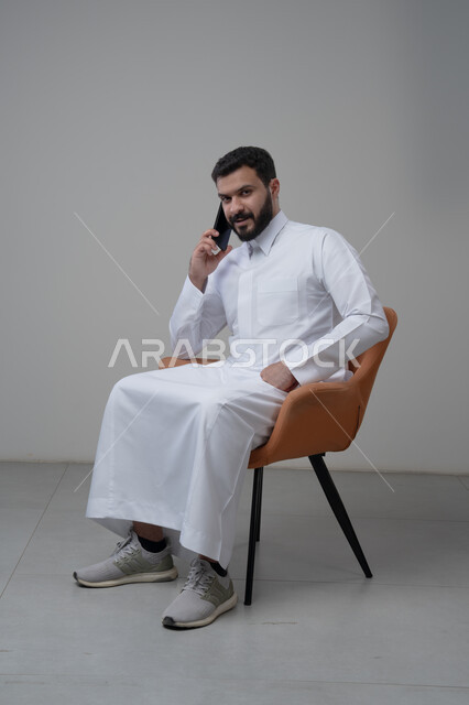 الرد على مكالمة هاتفية عبر الجوال، الدردشة والمحادثة باستخدام جهاز تقني حديث, بورتريه لرجل عربي خليجي سعودي يرتدي الثوب التقليدي يجلس على الكرسي يقوم باجراء اتصال هاتفي من خلال الهاتف المحمول، خلفية بيضاء