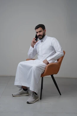 الرد على مكالمة هاتفية عبر الجوال، الدردشة والمحادثة باستخدام جهاز تقني حديث, بورتريه لرجل عربي خليجي سعودي يرتدي الثوب التقليدي يجلس على الكرسي يقوم باجراء اتصال هاتفي من خلال الهاتف المحمول، خلفية بيضاء
