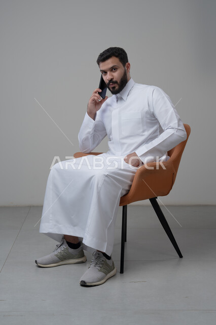 الرد على مكالمة هاتفية عبر الجوال، الدردشة والمحادثة باستخدام جهاز تقني حديث, بورتريه لرجل عربي خليجي سعودي يرتدي الثوب التقليدي يجلس على الكرسي يقوم باجراء اتصال هاتفي من خلال الهاتف المحمول، خلفية بيضاء