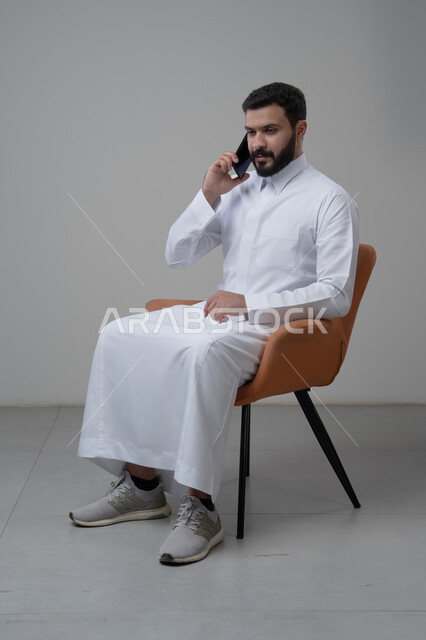الرد على مكالمة هاتفية عبر الجوال، الدردشة والمحادثة باستخدام جهاز تقني حديث, بورتريه لرجل عربي خليجي سعودي يرتدي الثوب التقليدي يجلس على الكرسي يقوم باجراء اتصال هاتفي من خلال الهاتف المحمول، خلفية بيضاء