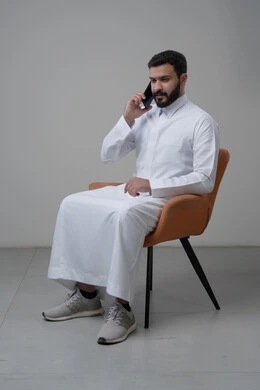 Answering a phone call via mobile phone, chatting and talking using a modern technical device, portrait of a Saudi Arabian Gulf man wearing traditional thobe sitting on a chair making a phone call via mobile phone, white background