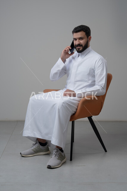 الرد على مكالمة هاتفية عبر الجوال، الدردشة والمحادثة باستخدام جهاز تقني حديث, بورتريه لرجل عربي خليجي سعودي يرتدي الثوب التقليدي يجلس على الكرسي يقوم باجراء اتصال هاتفي من خلال الهاتف المحمول، خلفية بيضاء