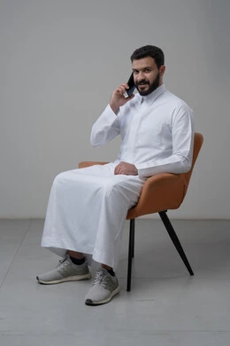 Answering a phone call via mobile phone, chatting and talking using a modern technical device, portrait of a Saudi Arabian Gulf man wearing traditional thobe sitting on a chair making a phone call via mobile phone, white background