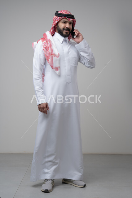 Chatting and conversation using a modern technical device, answering a phone call, portrait of a Saudi Arabian Gulf man wearing traditional thobe making a phone call through a mobile phone, full body image, white background