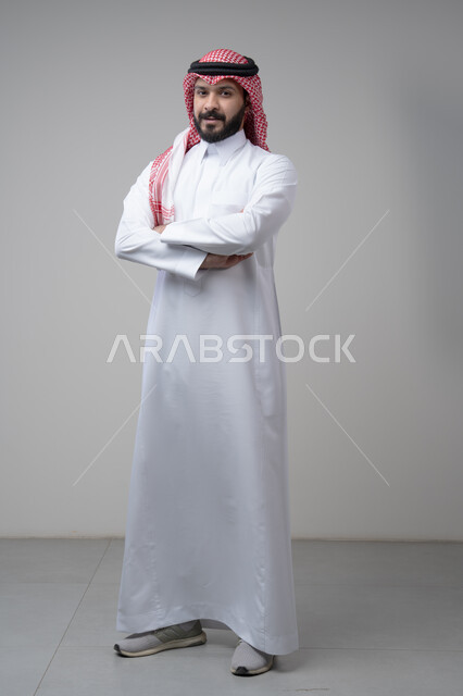  التقاط صور شخصية للوثائق الرسمية، الاعتناء والاهتمام بالمظهر الخارجي، مفهوم الاناقة والرجولة، بورتريه لشاب عربي خليجي سعودي يرتدي الثوب التقليدي يقف بأيدي متقاطعة ينظر الى الكاميرا بتعابير السرور والثقة بالنفس، الجسم بالطول الكامل، خلفية بيضاء