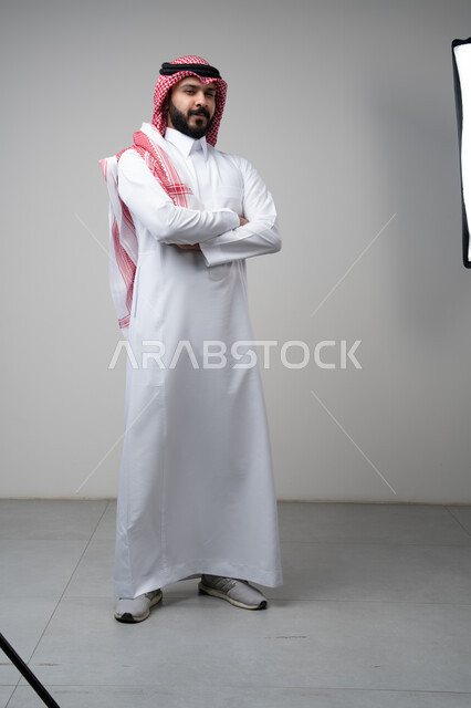  التقاط صور شخصية للوثائق الرسمية، الاعتناء والاهتمام بالمظهر الخارجي، مفهوم الاناقة والرجولة، بورتريه لشاب عربي خليجي سعودي يرتدي الثوب التقليدي يقف بأيدي متقاطعة ينظر الى الكاميرا بتعابير السرور والثقة بالنفس، الجسم بالطول الكامل، خلفية بيضاء