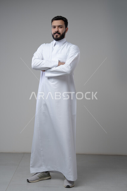  الاعتناء والاهتمام بالمظهر الخارجي، التقاط صور شخصية للوثائق الرسمية، مفهوم الاناقة والرجولة، بورتريه لشاب عربي خليجي سعودي يرتدي الثوب التقليدي يقف بأيدي متقاطعة ينظر الى الكاميرا بتعابير السرور والثقة بالنفس، الجسم بالطول الكامل، خلفية بيضاء
