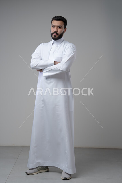  الاعتناء والاهتمام بالمظهر الخارجي، التقاط صور شخصية للوثائق الرسمية، مفهوم الاناقة والرجولة، بورتريه لشاب عربي خليجي سعودي يرتدي الثوب التقليدي يقف بأيدي متقاطعة ينظر الى الكاميرا بتعابير السرور والثقة بالنفس، الجسم بالطول الكامل، خلفية بيضاء