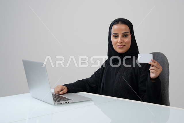 عرض موك اب ابيض صغير، وظيفة ومهنة نسائية مكتبية سعودية، ادارة وانجاز المهام باستخدام جهاز تقني حديث ومتطور, بورتريه لإمرأة عربية خليجية سعودية مبتسمة ترتدي الحجاب والعباءة السوداء تمسك بيدها بطاقة تعريفية تعمل على الحاسب المحمول, خلفية رمادية