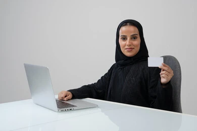 Small white mockup, Saudi female office job and profession, managing and completing tasks using a modern and advanced technical device, portrait of a smiling Saudi Arabian Gulf woman wearing a hijab and black abaya holding an ID card in her hand working on a laptop, gray background