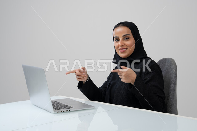 وظيفة ومهنة نسائية مكتبية سعودية، ادارة وانجاز المهام باستخدام جهاز تقني حديث ومتطور, بورتريه لإمرأة عربية خليجية سعودية ترتدي الحجاب والعباءة السوداء تشير الى الحاسوب المحمول تعمل في مقر الشركة, خلفية رمادية