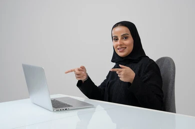 Saudi female office job and profession, managing and completing tasks using a modern and advanced technical device, portrait of a Saudi Arabian Gulf woman wearing a hijab and black abaya pointing at a laptop working at the company headquarters, gray background